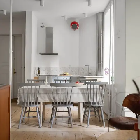 A Scandinavian dining nook with a grey-painted spindle-back dining set on light wood flooring, opening to a marble-backsplash kitchen counter behind.