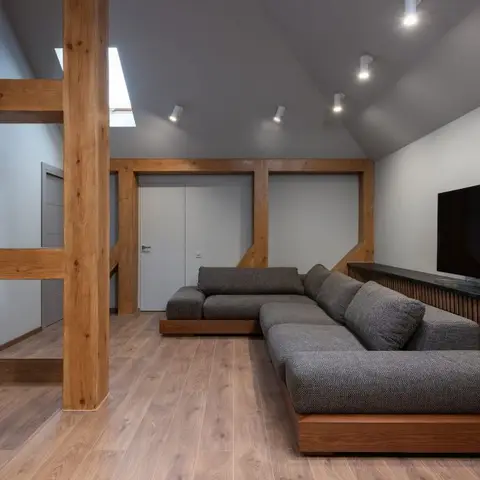 A sloped-ceiling loft living room with exposed wood beam columns, a wall-mounted TV over a fluted wood console, and a grey sectional sofa on wood flooring.
