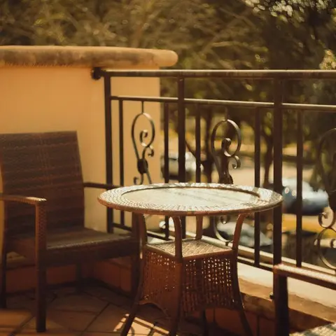 A small balcony corner with a wicker chair and round table beside an ornate scrollwork iron railing, lit by warm afternoon sun.