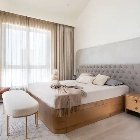 A small dog sits on a king bed with a tufted grey headboard against a curved plaster wall, tan leather swivel chair in the corner