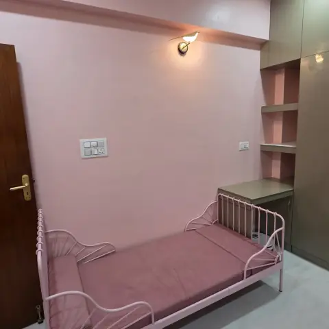 A small dusty-pink room with an extendable pink metal-frame child's bed and a thin pink mattress against the long wall