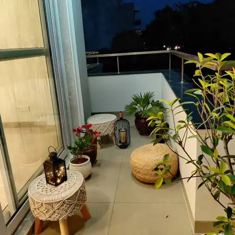 A small home balcony styled with macrame-topped stools, a braided jute pouf, potted plants, and string-light lanterns at night.
