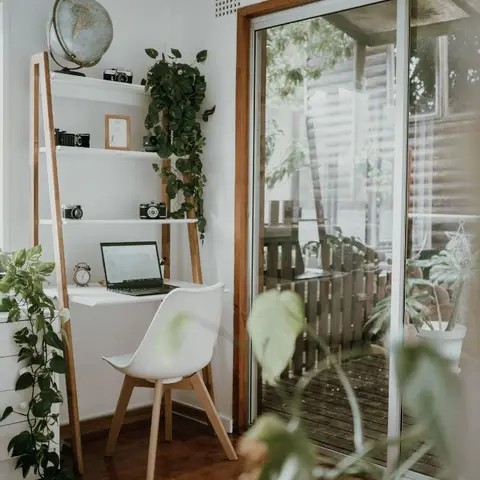 A small home-office nook with a white ladder desk and laptop beside a sliding glass door, framed by trailing pothos plants and vintage camera curios.