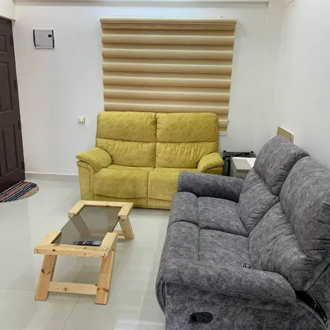 A small living room with a mustard-yellow two-seater and a grey fabric sofa around a raw pine coffee table with a glass top, beneath a zebra roller blind