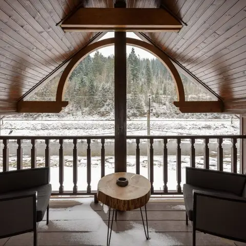 A snow-covered timber-framed balcony with two upholstered lounge chairs flanking a live-edge wood slab side table beneath an arched wooden gable.