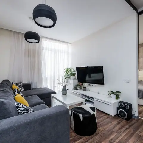 A studio living area with a grey sectional sofa, black drum pendant lights, sheer curtains, and a white TV console