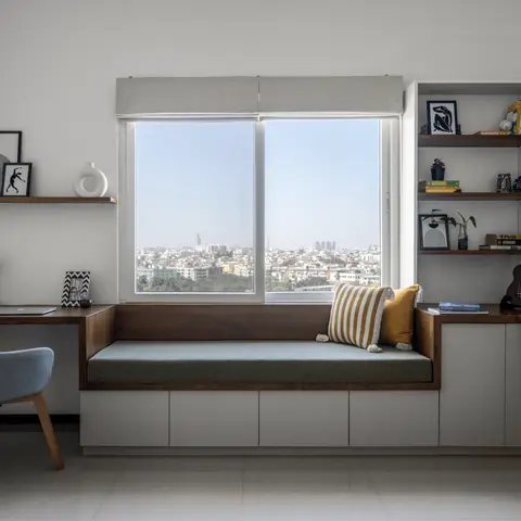 A study nook with a built-in wood window seat with storage drawers below, flanking open shelving styled with books and framed art, overlooking a city skyline.