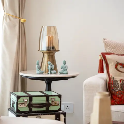 A styled living room corner with a round side table holding a brass hurricane lamp and jade Buddha figurines atop stacked vintage-style trunks.