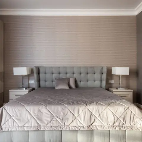 A symmetrical master bedroom with a grey tufted wingback headboard, matching nightstand lamps and a full-height wardrobe wall against textured wallpaper.