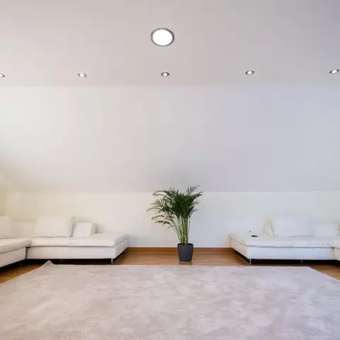 A symmetrical white sectional lounge under a sloped attic ceiling, with a single potted areca palm centered against the back wall and a pale rug on wood