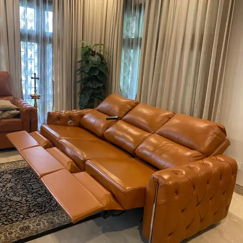 A tan tufted-leather reclining sofa shown fully extended, in a living room with floor-to-ceiling patterned sheer curtains, a traditional rug