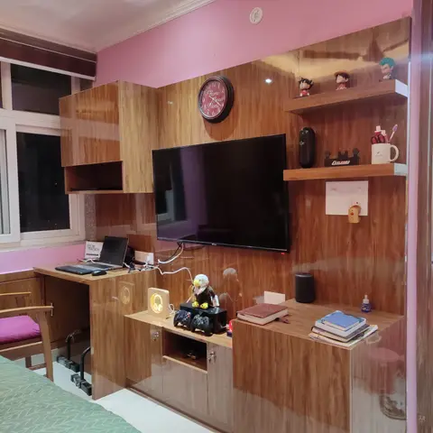 A teenager's pink-walled bedroom with a wood-laminate study-and-TV unit cluttered with anime figurines and a game console