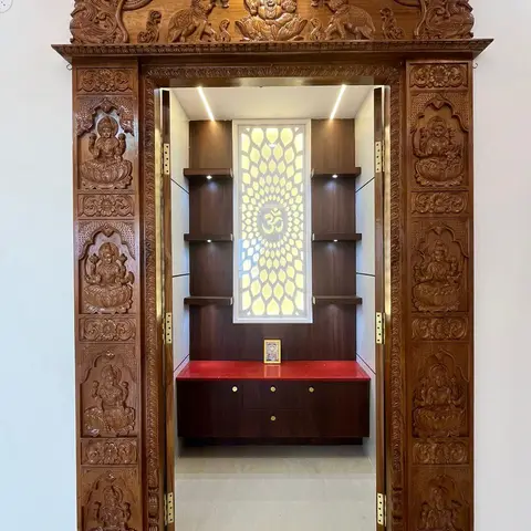 A traditional hand-carved wood pooja unit with Ganesh and Lakshmi relief door panels, a backlit floral Om jaali panel, and a red stone-topped cabinet inside.