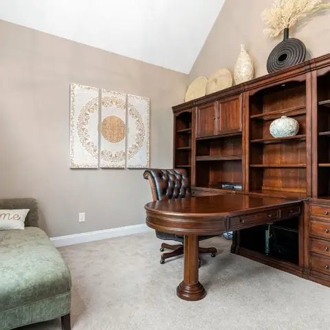 A traditional home study with a dark wood built-in bookcase-and-desk unit, a leather desk chair, and a green velvet chaise beneath a triptych wall art piece.