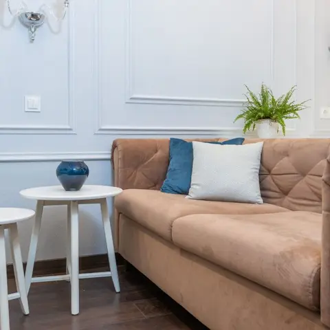 A tufted tan chesterfield-style sofa with blue velvet cushions sits against a white wainscot-panelled wall flanked by crystal candle wall sconces.