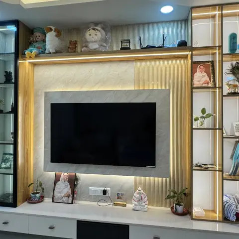 A TV feature wall with marble and fluted gold-trimmed panelling, flanked by open display shelving styled with family photos, plants, and personal items.