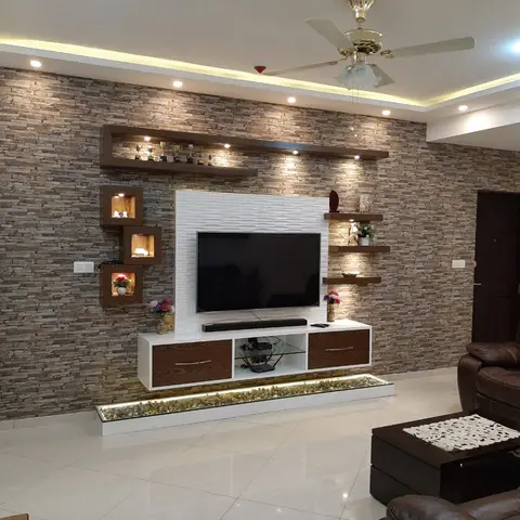 A TV unit with a white 3D-textured panel and floating display cubbies sits against a stacked stone-clad wall, lit by a pebble-filled light trough at its base.