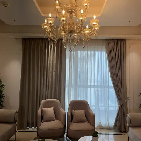 A twelve-arm amber crystal chandelier hangs low over two taupe tub chairs set against full-height velvet drapes with white sheers