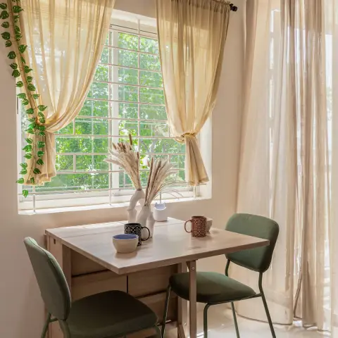 A two-seater light-wood drop-leaf table set into the corner below a grilled window, with two sage-green upholstered chairs on slim green legs