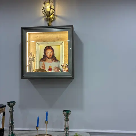 A wall-mounted illuminated Christian prayer shrine with a framed image of Jesus and religious figurines, above a marble console styled with brass candlesticks.