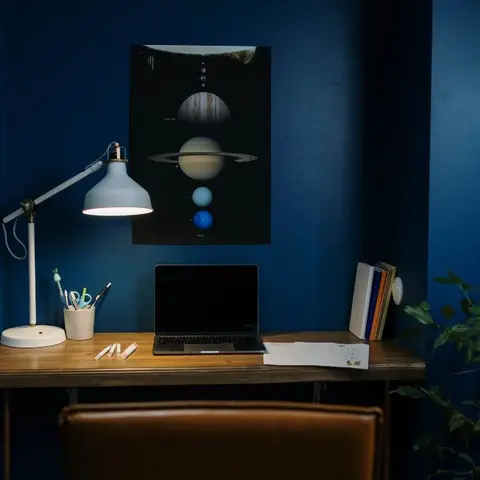 A wall-mounted wood study desk against a deep navy accent wall, with a framed solar-system poster and an adjustable desk lamp.