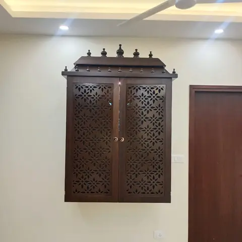 A wall-mounted wooden mandir unit with intricate floral jaali-cut doors and temple-style finials, set against a plain wall below a cove-lit ceiling