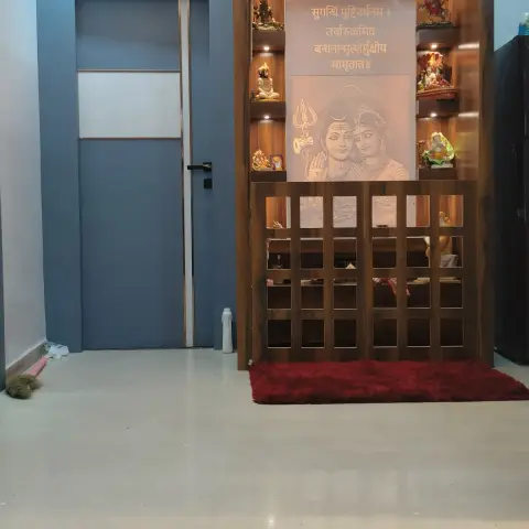 A wall-mounted wooden pooja unit with a jaali lattice lower cabinet, backlit deity niches, and a Shiva-Parvati motif panel