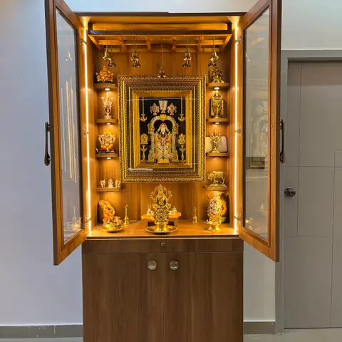 A wall-mounted wooden pooja unit with glass display doors, integrated LED shelf lighting, hanging brass bells