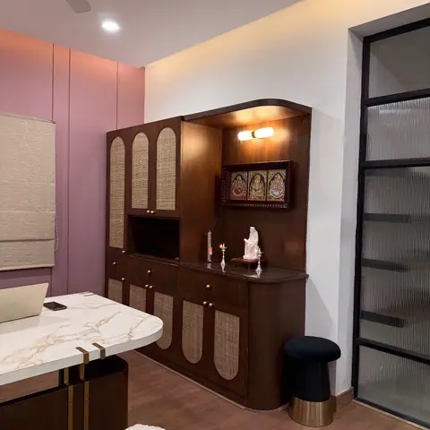A walnut-veneer pooja unit with cane-panel doors and a spotlit idol niche, beside a marble-top dining table and pink fluted wall panelling.