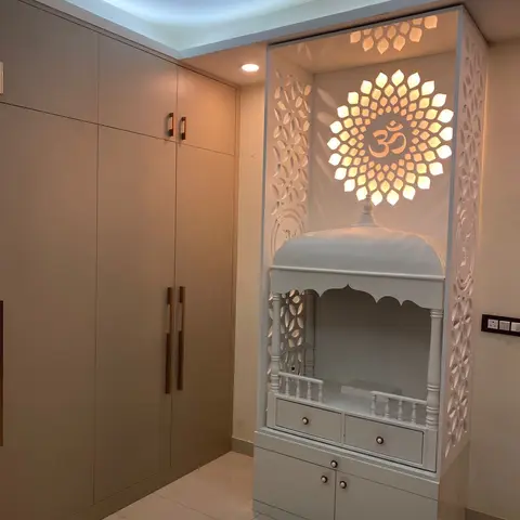 A white carved pooja unit with a backlit lotus-and-Om jaali panel and temple-arch canopy, set beneath a cove-lit ceiling beside a wardrobe wall.