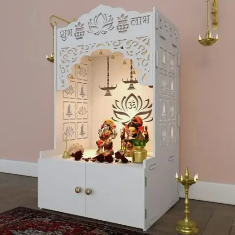 A white carved-wood pooja mandir unit with cabinet storage below, an illuminated idol niche, and hanging brass diyas, set against a blush pink wall.
