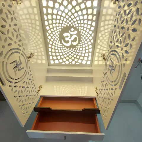 A white CNC-cut jaali pooja unit with a backlit Om motif and swastik carvings, its base drawer pulled open.
