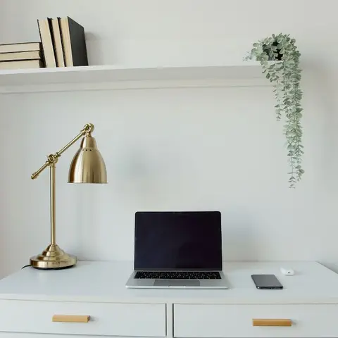 A white desk corner with a brass adjustable desk lamp, laptop, and a floating shelf of books above a trailing plant against a mustard curtain