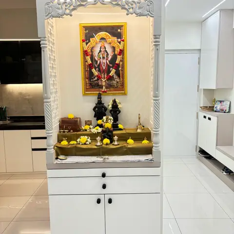 A white freestanding mandir with a carved fretwork pelmet and perforated jaali side panels, standing between the kitchen counter and the entrance shoe unit