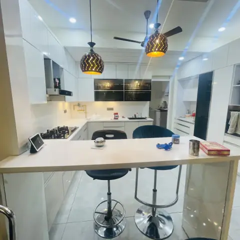 A white gloss U-shaped kitchen with a wood-topped breakfast counter, two patterned pendant lights, black bar stools, and integrated black gloss tall units.