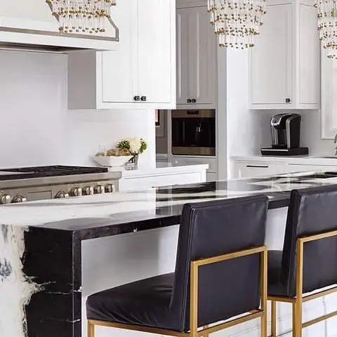 A white kitchen with a dramatic black-and-white marble waterfall island, gold beaded chandeliers overhead, and black leather bar stools with brass frames.