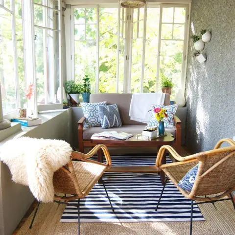 A window-lined sunroom with rattan armchairs, a striped dhurrie layered over a jute rug, and a wicker drum pendant light.
