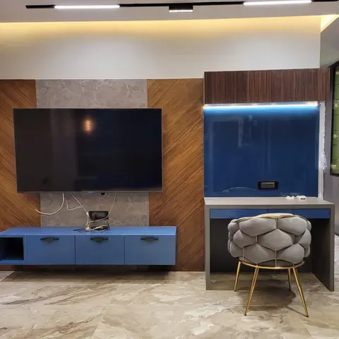 A wood-and-stone panelled TV wall with a floating blue console sits beside a glossy blue study desk and a honeycomb-tufted gold-leg stool.
