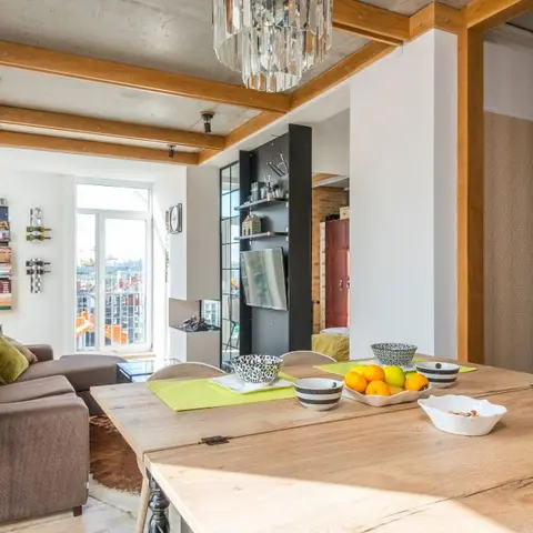A wood dining table set with fruit bowls in an open-plan apartment with an exposed brick wall, timber ceiling beams