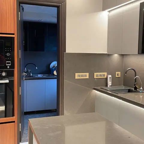 A wood-finish tall kitchen unit with a built-in oven and microwave stands beside grey stone countertops and backsplash with a black gooseneck faucet.