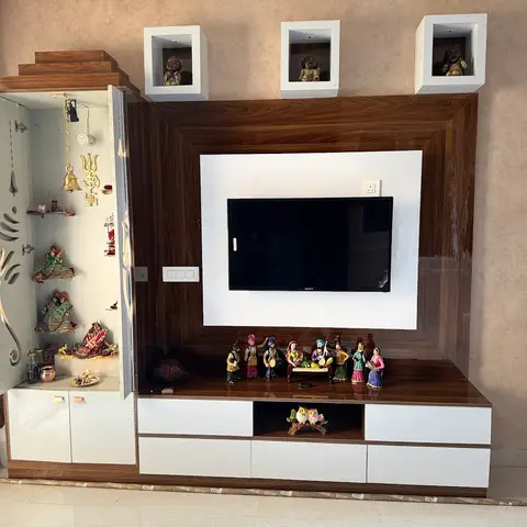 A wood-finish TV unit wall with an integrated pooja cabinet on one side and lit display niches holding figurines above.