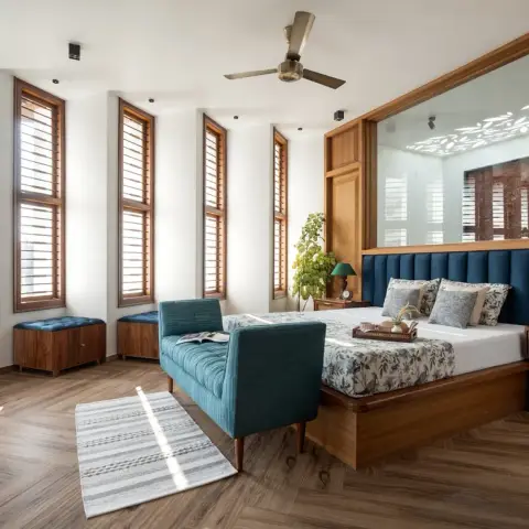 A wood-floored bedroom with a tall louvred window wall, a backlit jaali screen behind a teal channel-tufted headboard