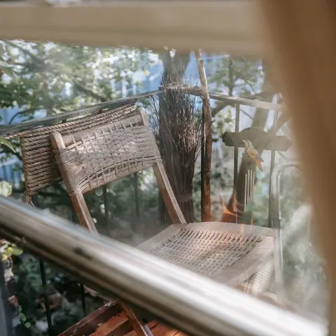 A woven cane balcony chair with a dried-reed bundle and small bird ornament, viewed through a window pane fringed by potted greenery.
