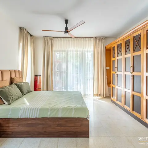 Airy bedroom where a full-wall teak-framed wardrobe with X-braced grey infill panels faces a low dark-wood bed with a tan channel-tufted headboard and a