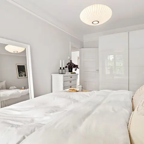 All-white bedroom with a high-gloss sliding-door wardrobe, a full-length leaning mirror, and a paper globe pendant light