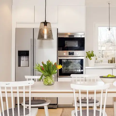 All-white kitchen with a built-in double oven column beside an American fridge, smoked-glass pendants over a pale wood table with spindle-back chairs
