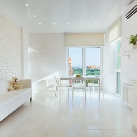 All-white open-plan studio with a whitewashed brick accent wall behind a leather sofa, a glass dining table by the balcony door