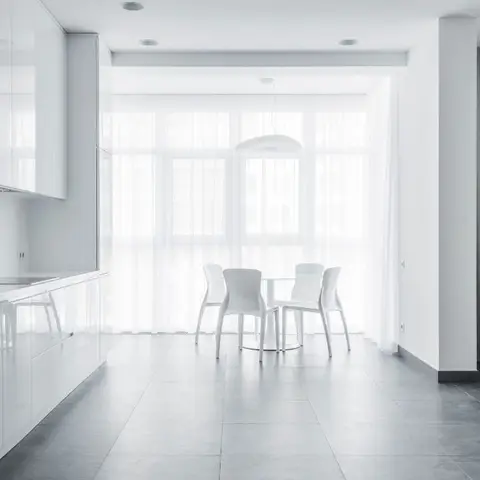 An all-white open-plan kitchen, dining and living space with sheer floor-to-ceiling curtains, a round glass dining table, and dark grey floor tiles.