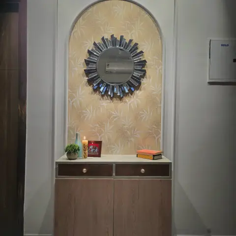An arched wallpapered niche with a sunburst mirror above a two-drawer wood-veneer entryway console styled with a vase, candle and stacked books.