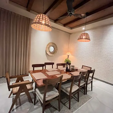 An eight-seat dining table with cane-back and Pierre Jeanneret-style chairs beneath woven basket pendant lights, exposed wood-beam ceiling